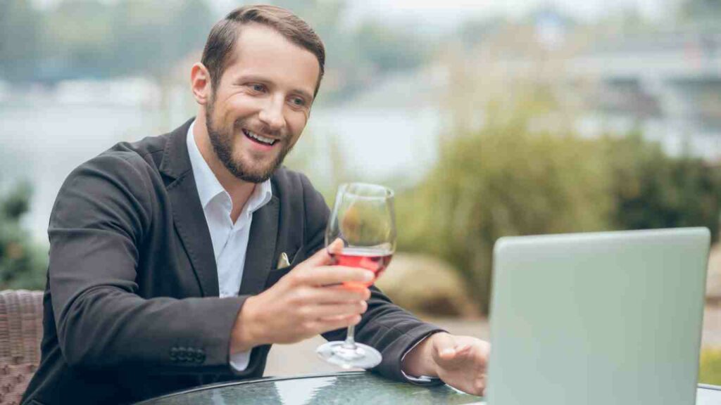 Online dating is about sending the right message (man looking at laptop and interacting with women online whilst having a glass of wine)